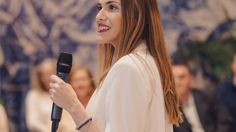 Junge Frau in einem weißen Blazer hält ein Mikrofon und spricht vor einem Publikum in einem modernen Veranstaltungsraum mit kunstvoller Wandgestaltung.