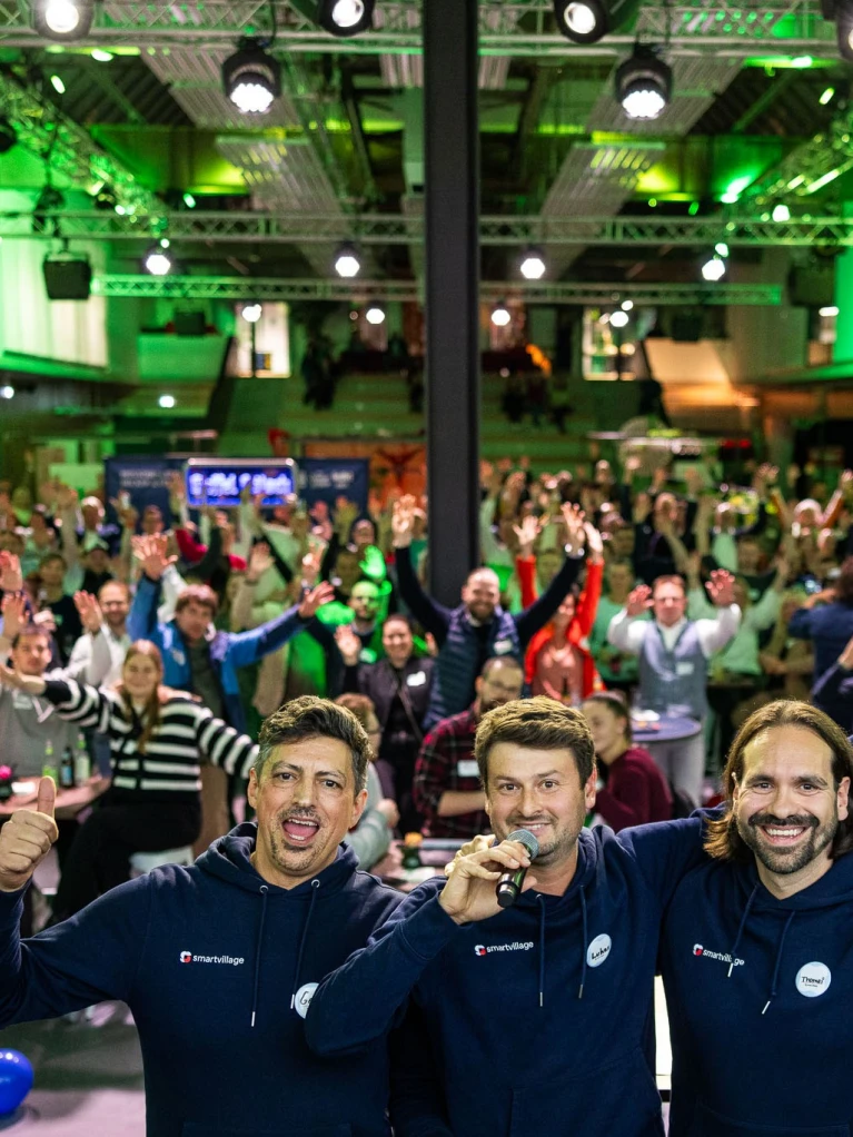 Drei Moderatoren auf einer Bühne mit Mikrofonen vor einer großen, begeisterten Menschenmenge bei einem Event in einer modern beleuchteten Veranstaltungshalle.