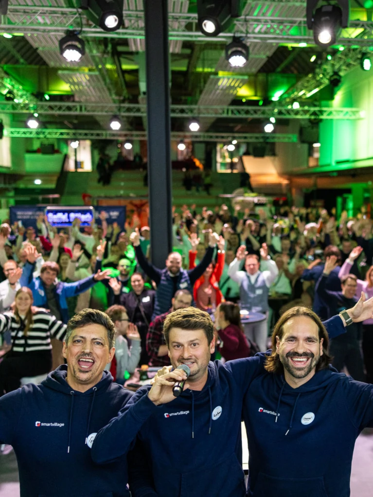 Drei Moderatoren auf einer Bühne mit Mikrofonen vor einer großen, begeisterten Menschenmenge bei einem Event in einer modern beleuchteten Veranstaltungshalle.
