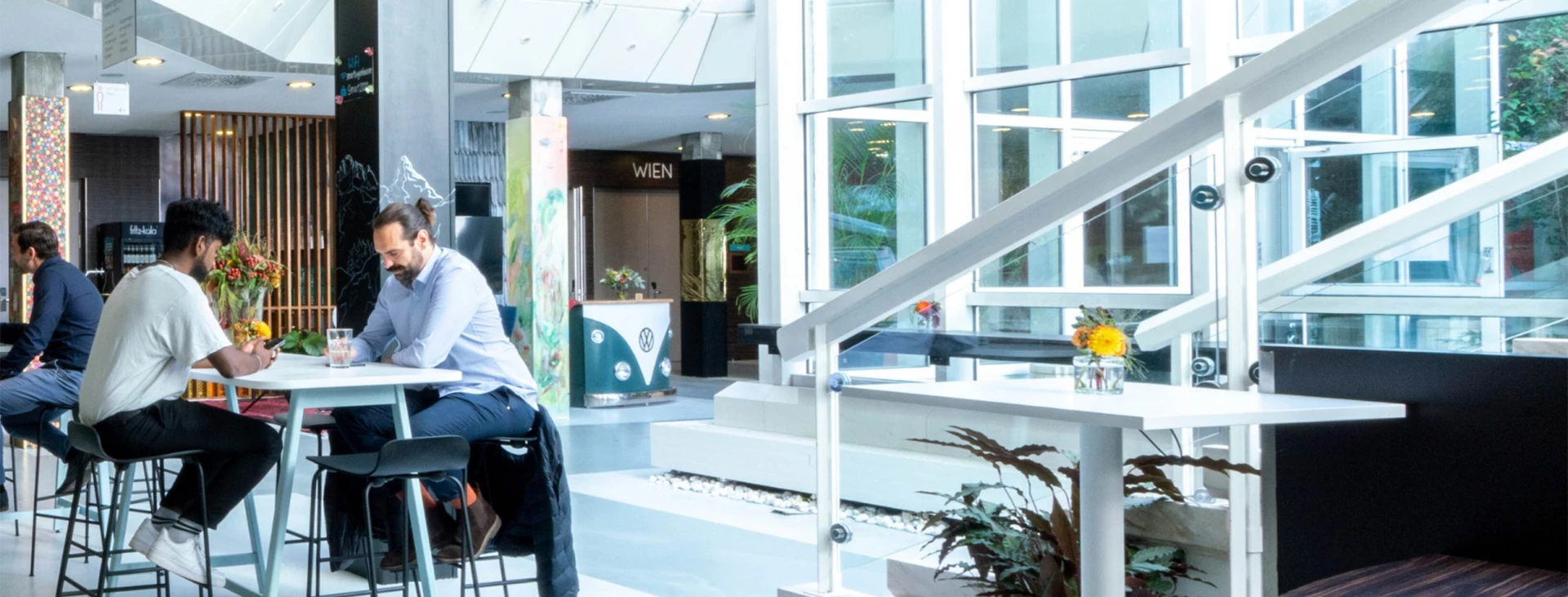 odernes Café oder Coworking-Space mit großen Fenstern, hohen Tischen und einer entspannten Atmosphäre. Zwei Männer unterhalten sich, während andere Gäste arbeiten.