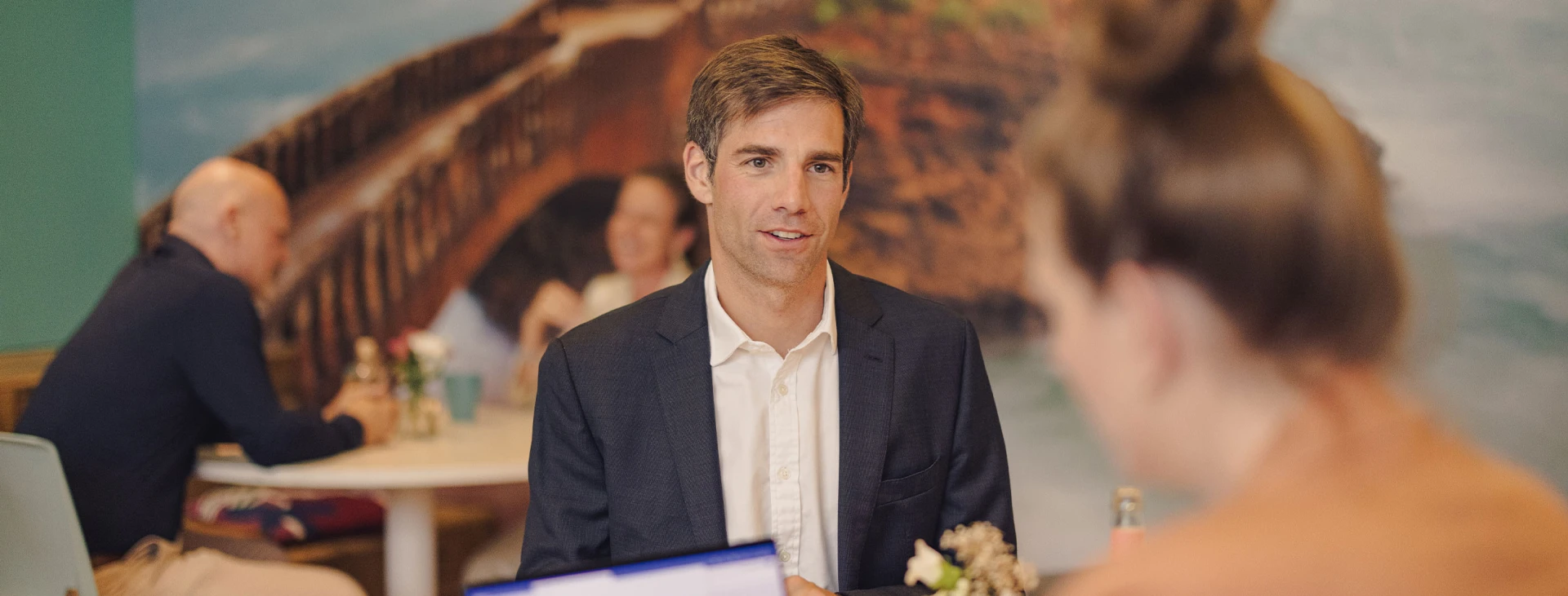 Geschäftsmann in Anzug im Gespräch mit einer Frau in einem modernen Café mit stilvollem Wanddesign. Weitere Gäste führen entspannte Unterhaltungen im Hintergrund.