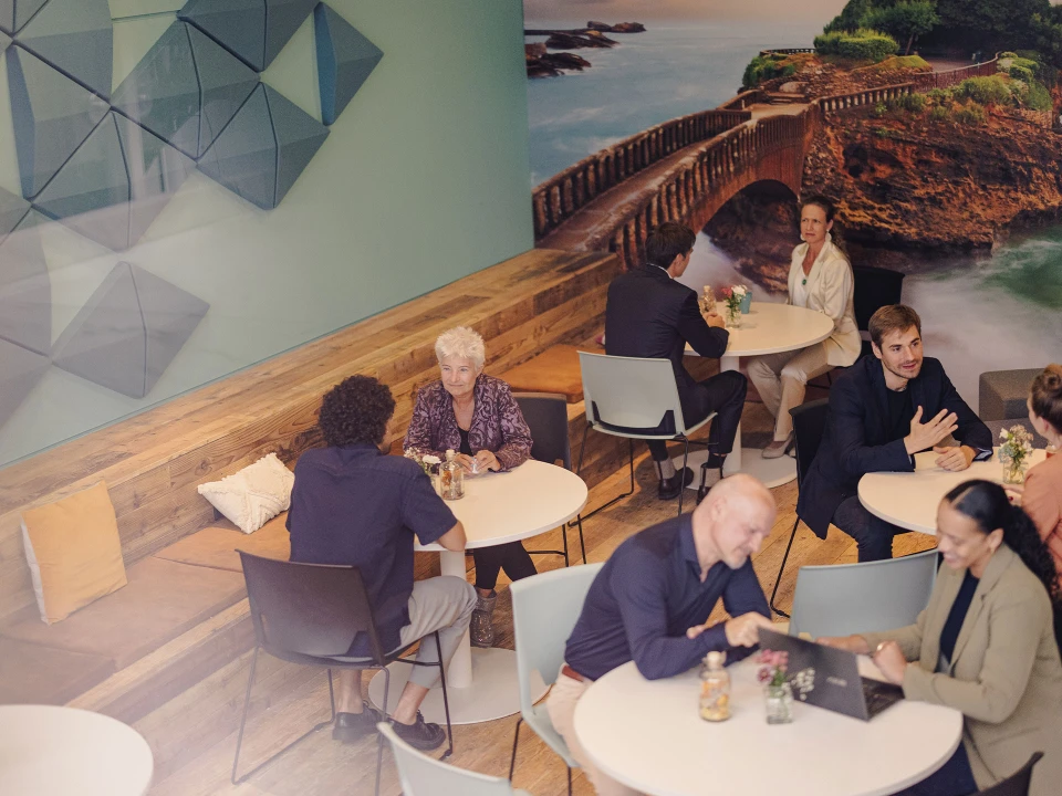 Gemütliches Café mit modernen Sitzgelegenheiten, Holzelementen und einer großen Wandgestaltung mit Naturmotiv. Gäste unterhalten sich oder arbeiten entspannt an Laptops.