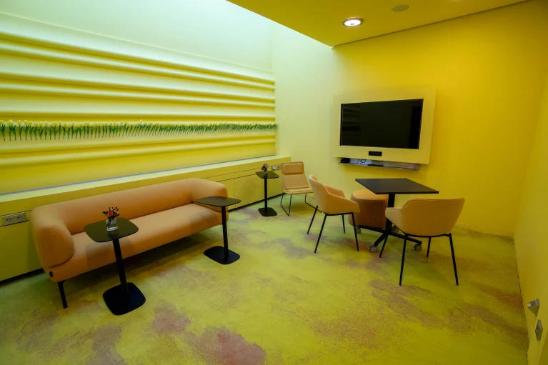 Design-Meetingraum mit einer gelben Lounge-Ecke, einem großen Wandbild aus künstlichen Tulpen, mehreren Sitzgelegenheiten und einem an der Wand montierten Fernseher – ideal für kreative Workshops im Smartvillage Bogenhausen