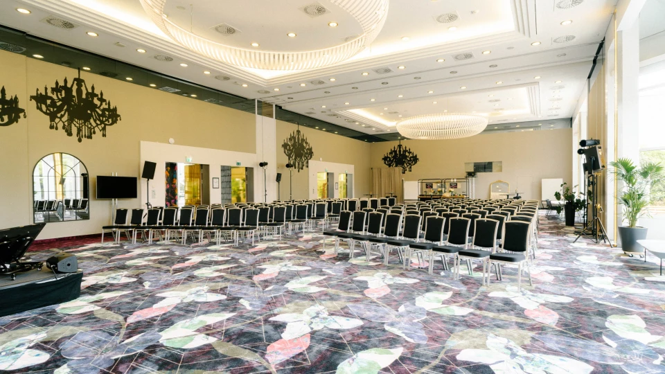 Moderner Veranstaltungssaal mit stilvoller Bestuhlung für Tagungen und Events. Hohe Decken mit Kronleuchtern, große Fenster für viel Tageslicht und ein farbenfroher Teppich schaffen eine elegante Atmosphäre