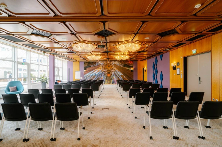 Modern eingerichteter Konferenzraum mit einer warmen Holzdecke, luxuriösen Kronleuchtern und einer beeindruckenden Wandgestaltung im futuristischen Stil. Die Reihen mobiler schwarzer Stühle sind auf eine Präsentationsfläche ausgerichtet, während große Fenster für viel Tageslicht sorgen. Der Raum verbindet klassisches Design mit innovativen Akzenten und eignet sich ideal für Tagungen, Workshops und Events