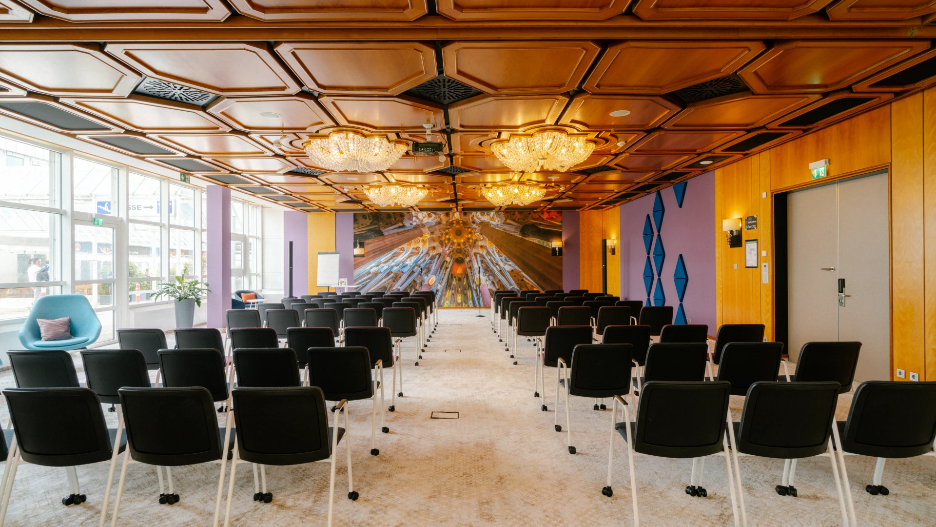Modern eingerichteter Konferenzraum mit einer warmen Holzdecke, luxuriösen Kronleuchtern und einer beeindruckenden Wandgestaltung im futuristischen Stil. Die Reihen mobiler schwarzer Stühle sind auf eine Präsentationsfläche ausgerichtet, während große Fenster für viel Tageslicht sorgen. Der Raum verbindet klassisches Design mit innovativen Akzenten und eignet sich ideal für Tagungen, Workshops und Events