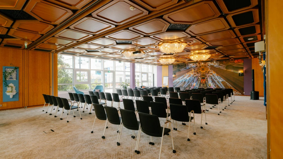 Stilvoller Veranstaltungsraum mit einer markanten Holzdecke, funkelnden Kronleuchtern und einer kreativen Wandgestaltung. Die Reihenbestuhlung mit schwarzen Stühlen auf Rollen ist perfekt für Konferenzen, Vorträge oder Seminare. Große Fenster an der Seite lassen viel Tageslicht herein und bieten einen Blick auf die grüne Außenanlage. Im hinteren Bereich sorgen gemütliche Sessel für eine entspannte Atmosphäre, während moderne Designelemente an den Wänden den Raum visuell aufwerten