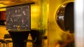 Champagner-Bar mit goldener Wandverkleidung, schwarzem Stehtisch und Hockern. Eine handbeschriftete Tafel mit der Aufschrift 'Champagne Bar' und dekorativen Illustrationen hängt an der Wand. Rechts spiegelt sich der Raum in einer runden, metallischen Öffnung.
