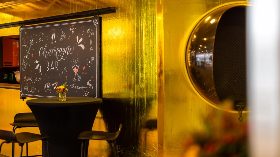 Champagner-Bar mit goldener Wandverkleidung, schwarzem Stehtisch und Hockern. Eine handbeschriftete Tafel mit der Aufschrift 'Champagne Bar' und dekorativen Illustrationen hängt an der Wand. Rechts spiegelt sich der Raum in einer runden, metallischen Öffnung.