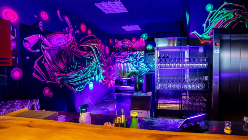 Moderne Bar mit neonfarbenem Graffiti an den Wänden, das eine dampfende Kaffeetasse und ein grimmiges Gesicht zeigt. Im Vordergrund eine Holztheke mit Flaschen und Barzubehör. Im Hintergrund ein Regal mit aufgereihten Gläsern, Weinflaschen und einer Metalltheke, beleuchtet von blauen und violetten Neonlichtern