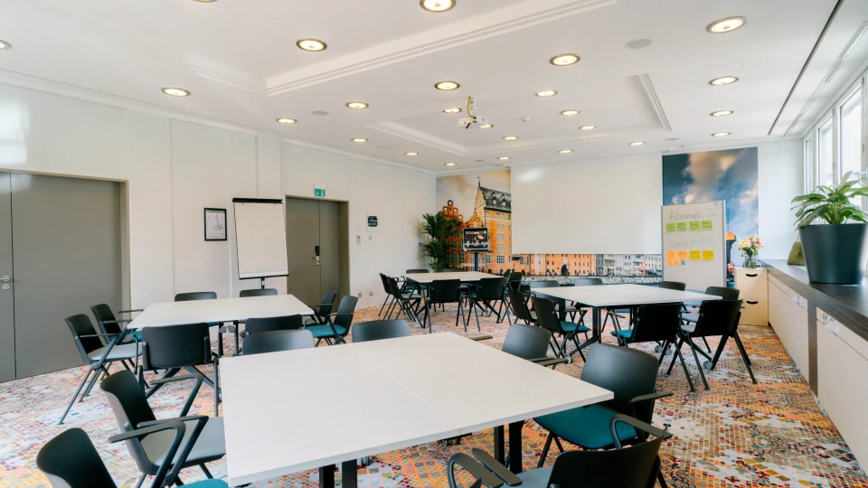 Heller und moderner Konferenzraum mit mehreren weißen Tischen und schwarzen Stühlen, angeordnet für Gruppenarbeit. An der Wand ein großes Stadtpanorama mit historischen Gebäuden. Eine Flipchart und ein Whiteboard mit Notizen stehen bereit. Große Fenster auf der rechten Seite lassen viel Tageslicht herein, während grüne Pflanzen den Raum ergänzen