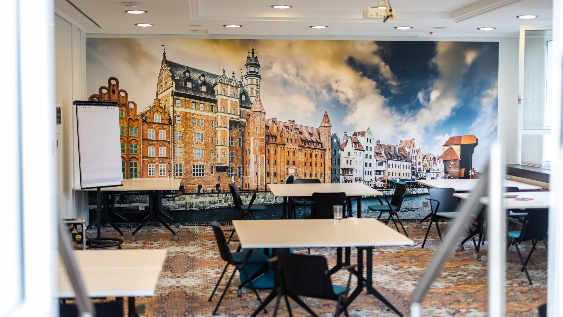 Moderner Seminarraum mit weißen Tischen und schwarzen Stühlen, bereit für Gruppenarbeiten. Eine Wand ist mit einem großen Fotomotiv einer historischen europäischen Stadt mit roten Backsteinhäusern und einem Fluss gestaltet. Eine Flipchart steht an der Seite, während helle Deckenstrahler den Raum ausleuchten