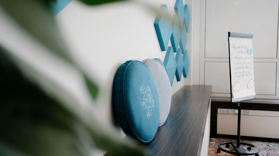 Detailaufnahme einer modernen Raumgestaltung mit zwei runden blauen Kissen auf einer Holzablage. Im Hintergrund eine Flipchart mit handgeschriebenem Text und geometrische, blau gepolsterte Wanddekorationen. Ein grünes Blatt im Vordergrund sorgt für eine weiche Unschärfe