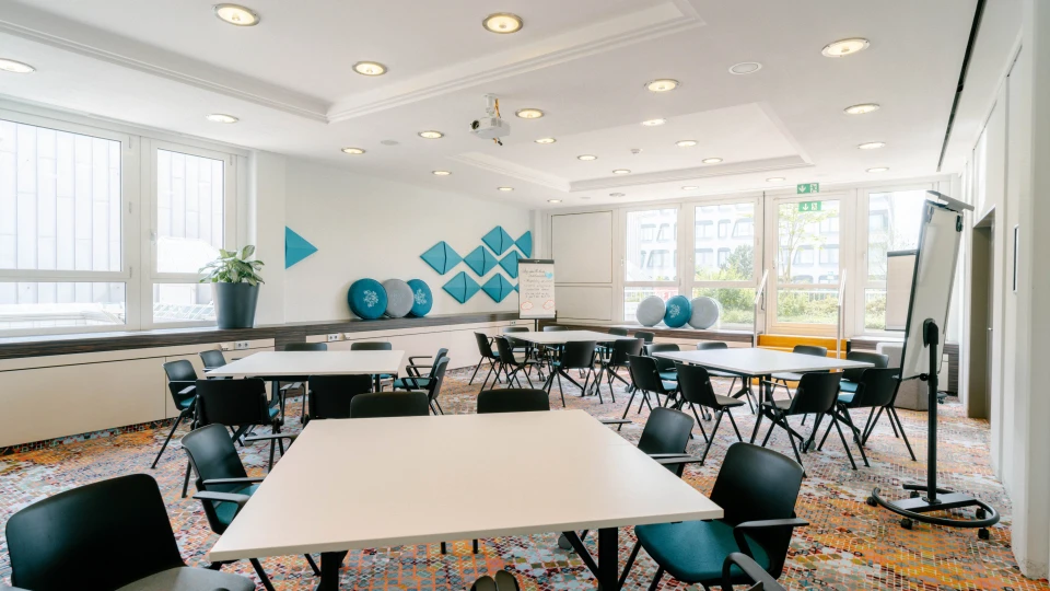 Heller und modern eingerichteter Konferenzraum mit mehreren weißen Tischen und schwarzen Stühlen. An den Wänden befinden sich blaue geometrische Polster und runde Dekokissen. Große Fenster lassen viel Tageslicht herein, während Flipcharts und eine Projektionsfläche für Präsentationen bereitstehen