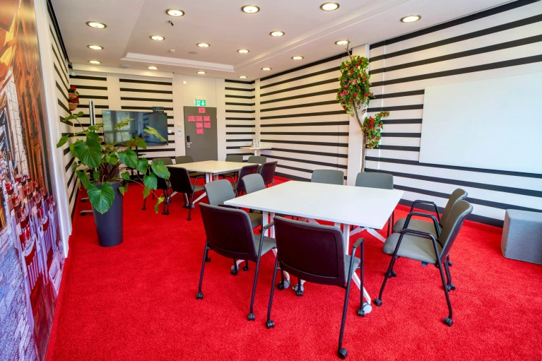 Moderner Konferenzraum mit schwarz-weiß gestreiften Wänden, roten Teppichen, weißen Tischen, Pflanzen und Wandtafel.