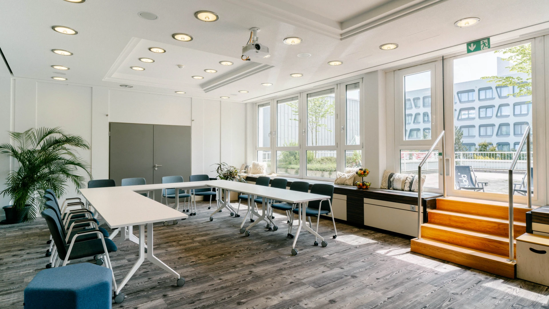 Heller Konferenzraum mit großen Fenstern, Holzfußboden, weißen Tischen, blauen Stühlen und Zugang zur Terrasse.