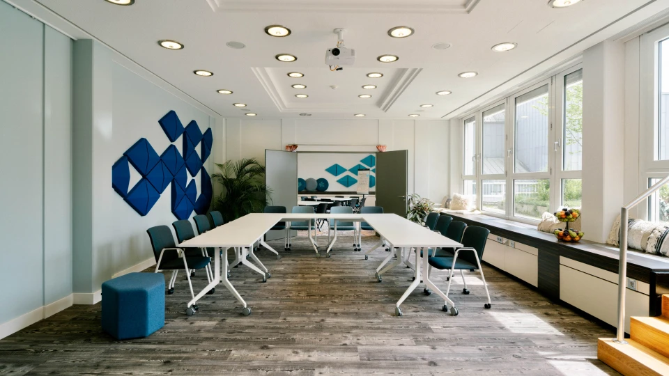 Heller Konferenzraum mit blauen Wandpaneelen, weißen Tischen auf Rollen, grauen Stühlen und großen Fenstern mit Sitzbank