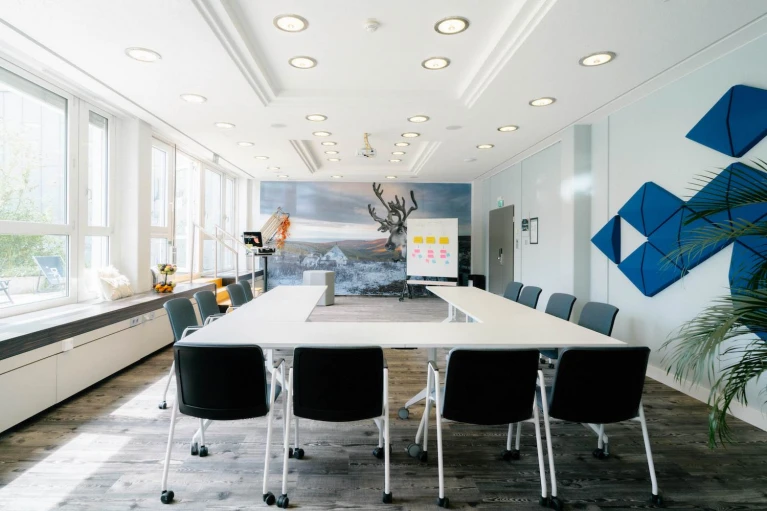 Moderner Konferenzraum mit U-förmiger Tischanordnung, großen Fenstern, Wandkunst mit Rentiermotiv und blauen Akustikpaneelen.