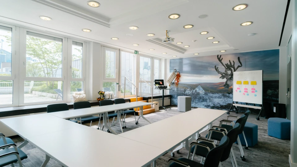 Heller Konferenzraum mit U-förmiger Tischanordnung, großen Fenstern, Rentier-Wandbild und Whiteboard mit Notizen.
