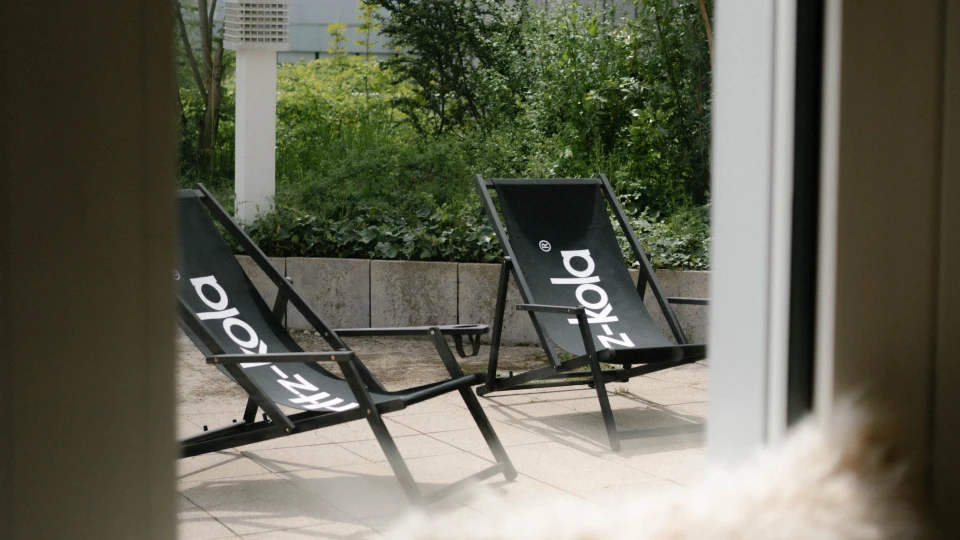 Blick durch ein Fenster auf eine Terrasse mit zwei schwarzen Liegestühlen, umgeben von grüner Bepflanzung.