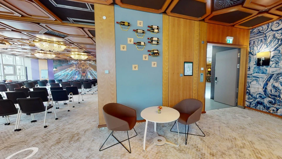 Lounge-Bereich mit zwei braunen Sesseln, rundem Tisch und Wanddekoration mit Weinflaschen, neben einem Konferenzraum