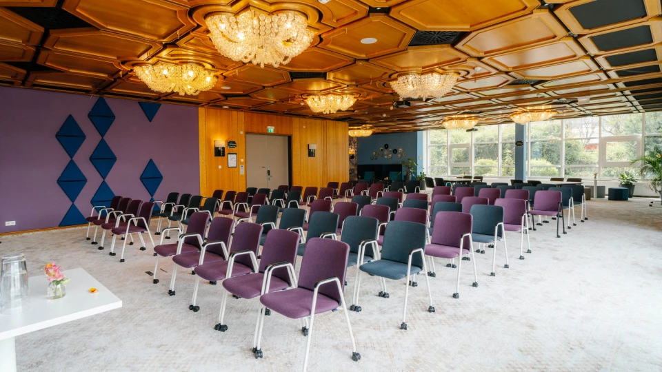 Großer Konferenzraum mit Reihenbestuhlung, Kristalllüstern, Holzkassettendecke und violett-blauen Wandakzenten