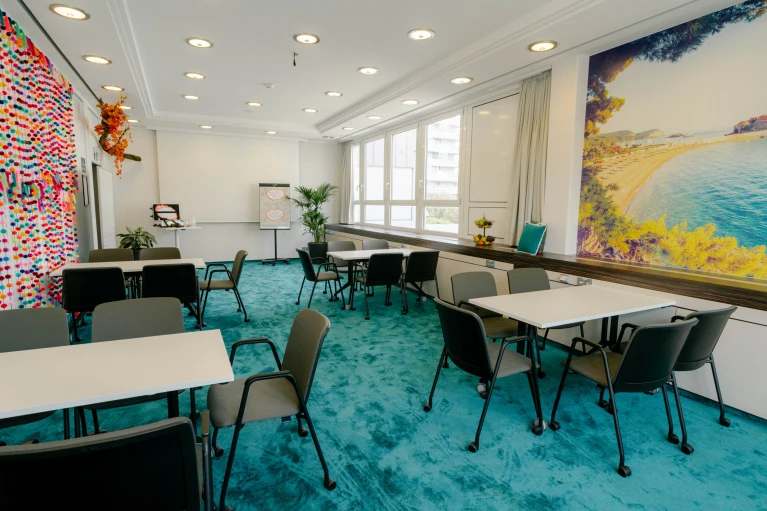 Heller Meetingraum mit türkisfarbenem Teppich, weißen Tischen und grauen Stühlen. Die Wand ist mit einem großen Strandmotiv sowie bunten Girlanden dekoriert, die eine kreative Atmosphäre schaffen