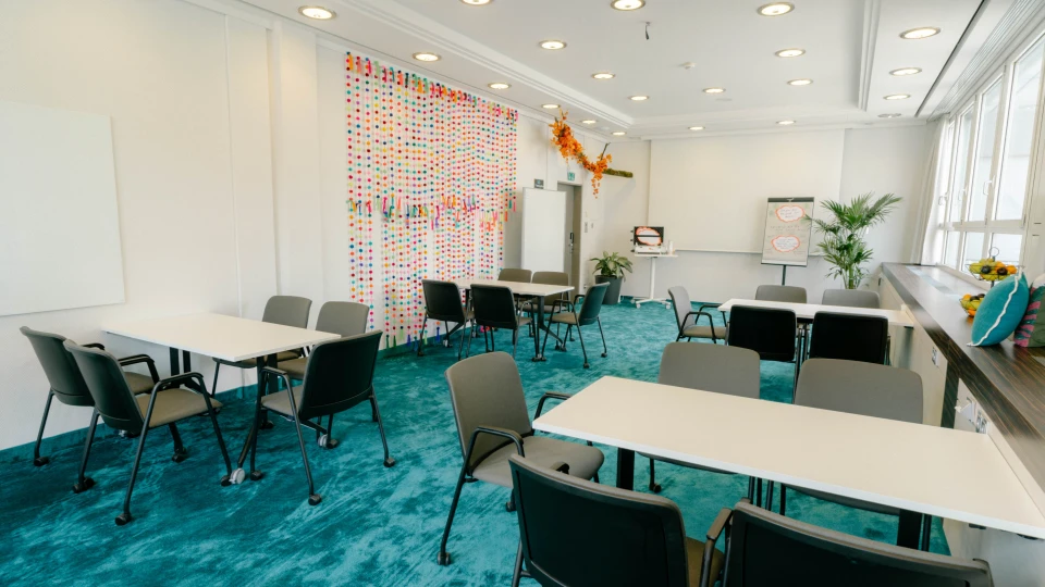 Kreativer Seminarraum mit blauem Teppich, bunten Pompon-Girlanden und flexibler Tischanordnung für Gruppenarbeiten
