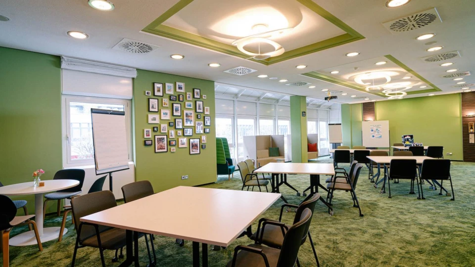 Meetingraum mit eckigen Tischen, türkisen Stühlen, grüner Wand und mediterranen Deko-Elementen.