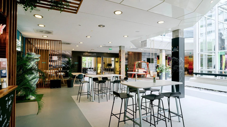 Heller Cafébereich mit Stehtischen, Hockern und Blick auf Besprechungsräume und Schaukelbank