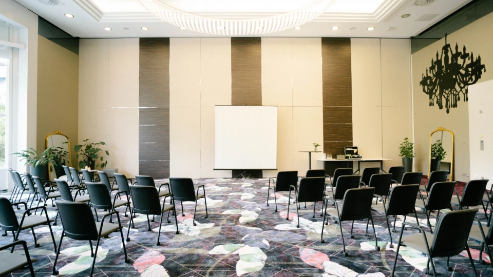 Symmetrischer Blick auf die Präsentationsfläche im Raum Versailles mit moderner Bestuhlung, Teppich mit Blattmuster und dekorativen Details.